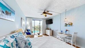 Un dormitorio con cama, ventilador de techo, televisión, mesa de comedor con sillas y vista a una piscina.