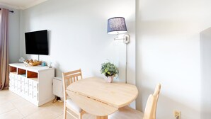 Un comedor con una mesa de madera, sillas, un armario blanco con cajones, un televisor montado en la pared y una lámpara de pie.