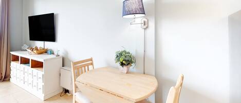 En matplats med ett träbord, stolar, ett vitt skåp med lådor, en tv som är monterad på väggen och en golvlampa.
