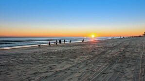 A beach with footprints and tire tracks, a sunset in the background, and people walking along the shore. - Blu 305 - Gulf-front condo with upgraded features - quick drive to entertainment (Fort Walton Beach)