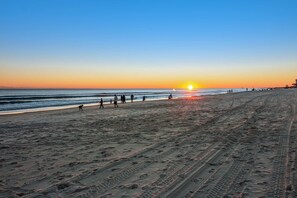 A beach with footprints and tire tracks, a sunset in the background, and people walking along the shore. - Blu 305 - Gulf-front condo with upgraded features - quick drive to entertainment (Fort Walton Beach)