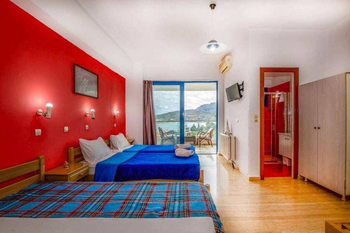 Una habitación con dos camas, una pared roja, un ventanal con vistas, un televisor y un armario.
