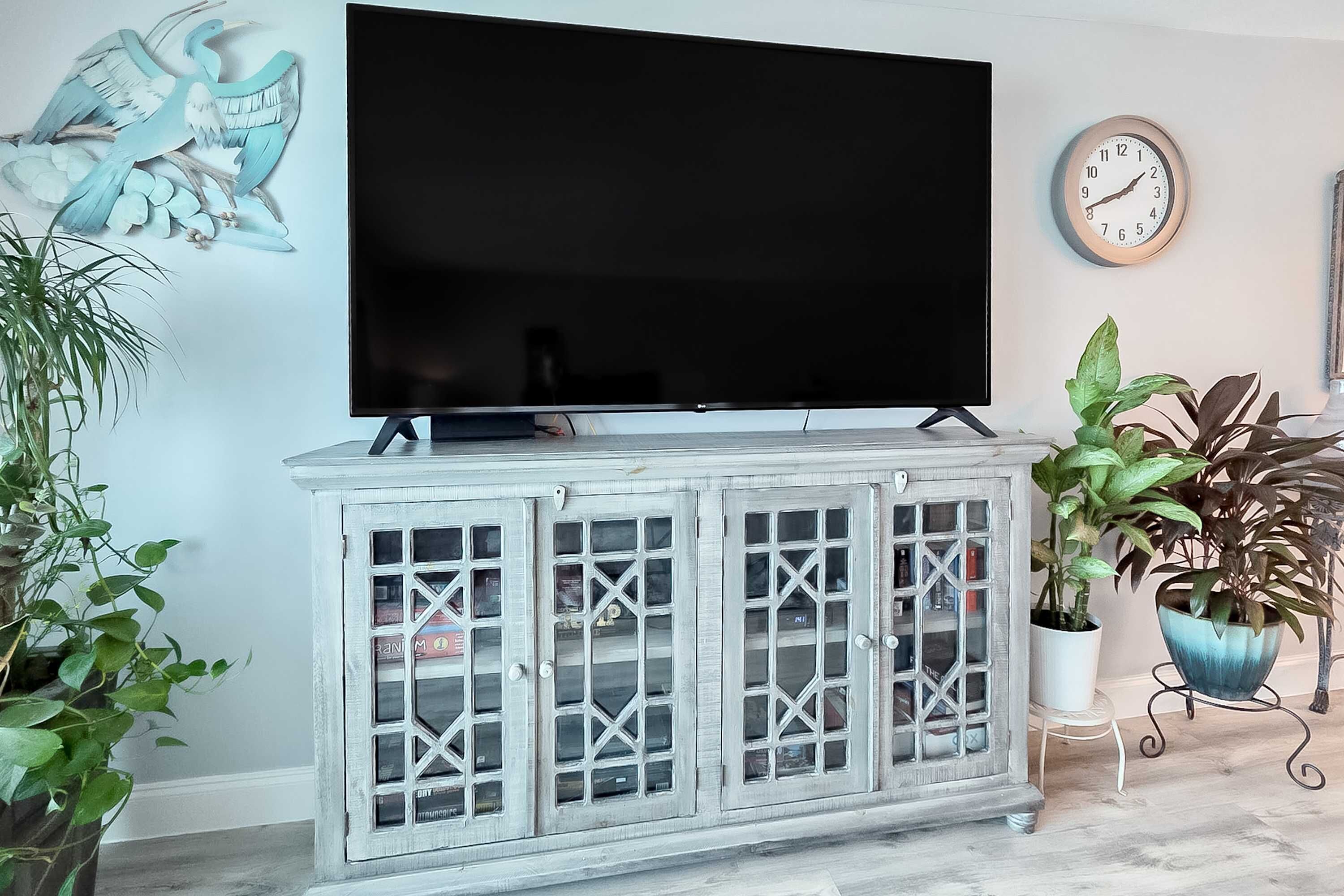 A living room with a flat-screen TV on a white cabinet, a clock on the wall, and potted plants.