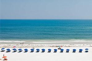 A beach with rows of blue and red beach chairs and umbrellas, a clear blue sea, and a sandy shore. - Fourth floor condo with partial Gulf views, WiFi, balcony, pool & tennis (Fort Walton Beach)