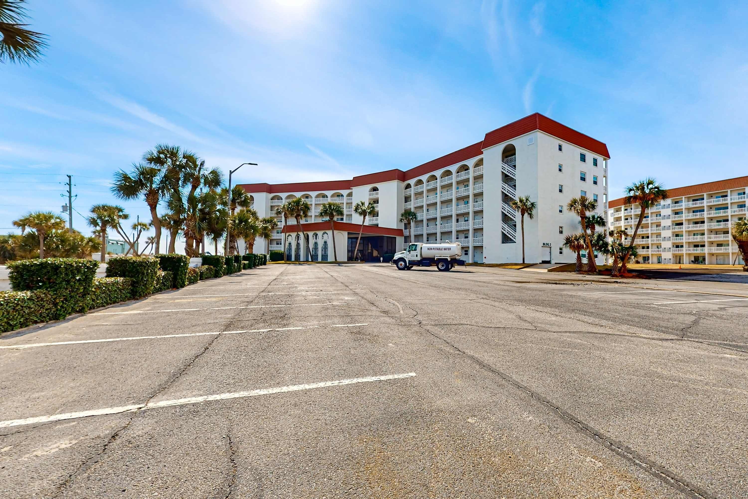 Un grand hôtel à plusieurs étages, avec un toit rouge, comprenant un parking où sont stationnées plusieurs voitures et bordé de palmiers.