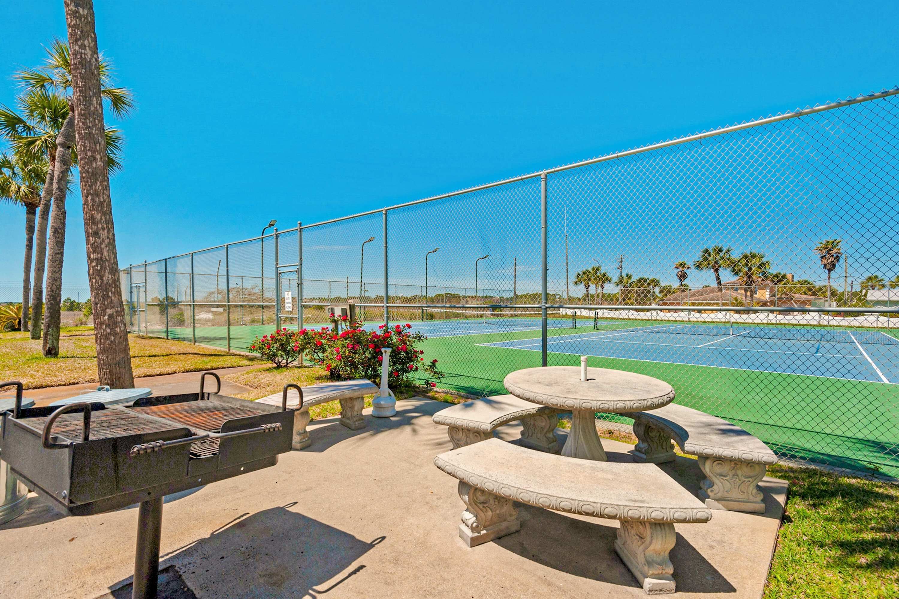An outdoor patio area with a barbecue grill, stone tables, and a tennis court in the background.