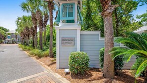 Una entrada con reja y un cartel que dice "Old Florida Cottages", rodeada de palmeras y un camino pavimentado.