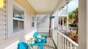 Un porche techado con sillas Adirondack azules, barandal blanco y vista a una zona residencial con cerca blanca y palmeras.