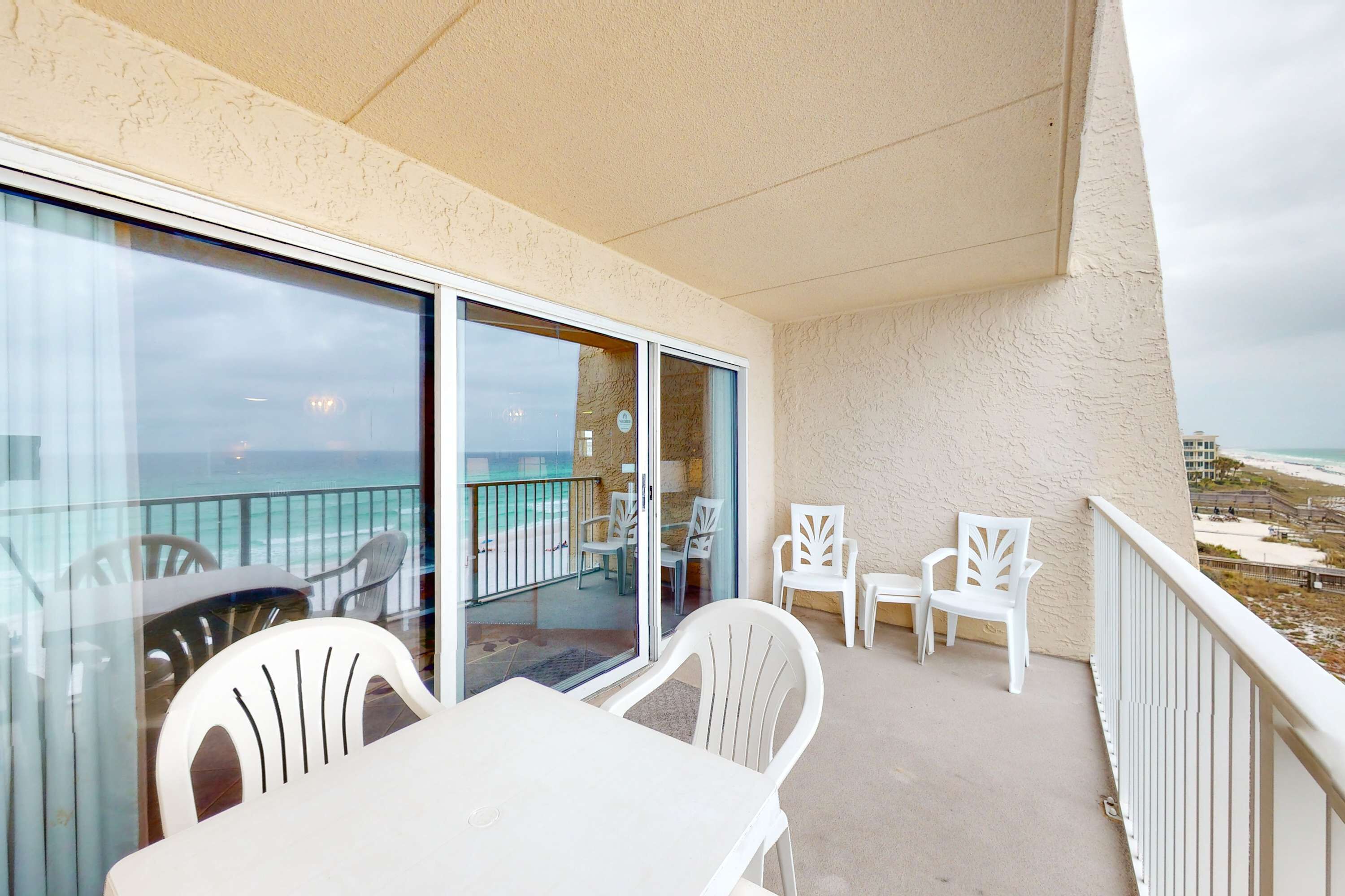 A balcony with white plastic chairs and a table, overlooking a beach and ocean.