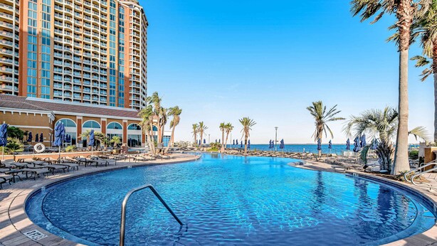 A large outdoor swimming pool with a surrounding lounge area, palm trees, and a tall hotel building in the background. - Waterfront condo with WiFi, washer/dryer, pools, fitness room, & beach access (Pensacola Beach)