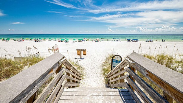 Ein Strand mit Holzstegen, die zum Ufer führen, strahlend blauem Himmel und einem Sandstrand mit Personen und Sonnenschirmen.