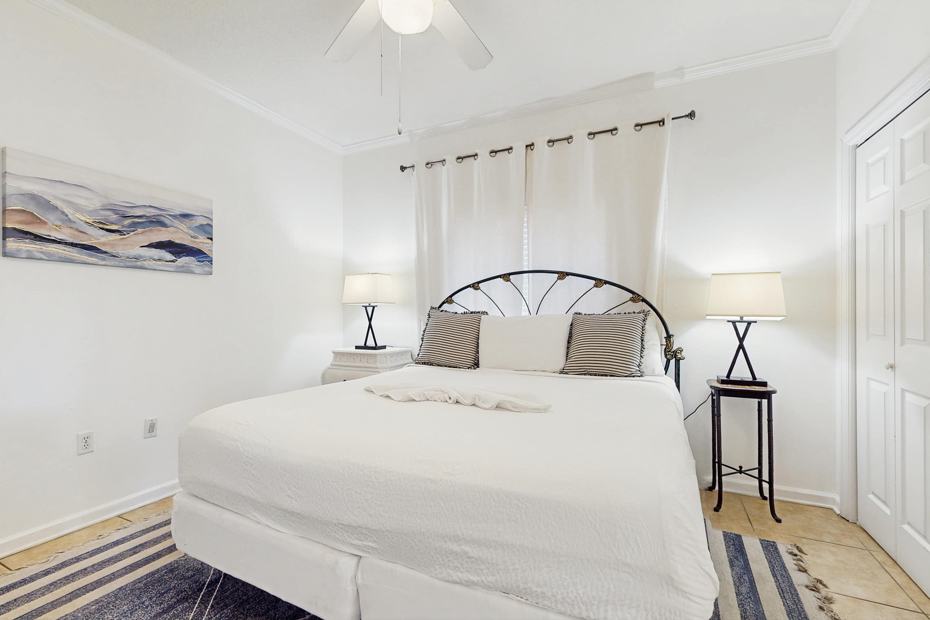 A bedroom with a bed, two bedside tables with lamps, a ceiling fan, a painting on the wall, and a door.
