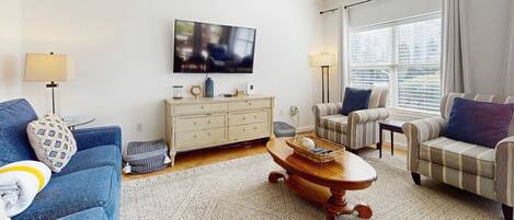 A living room with a blue sofa, a striped armchair, a wooden coffee table, a TV on the wall, a white cabinet, and a window with curtains.