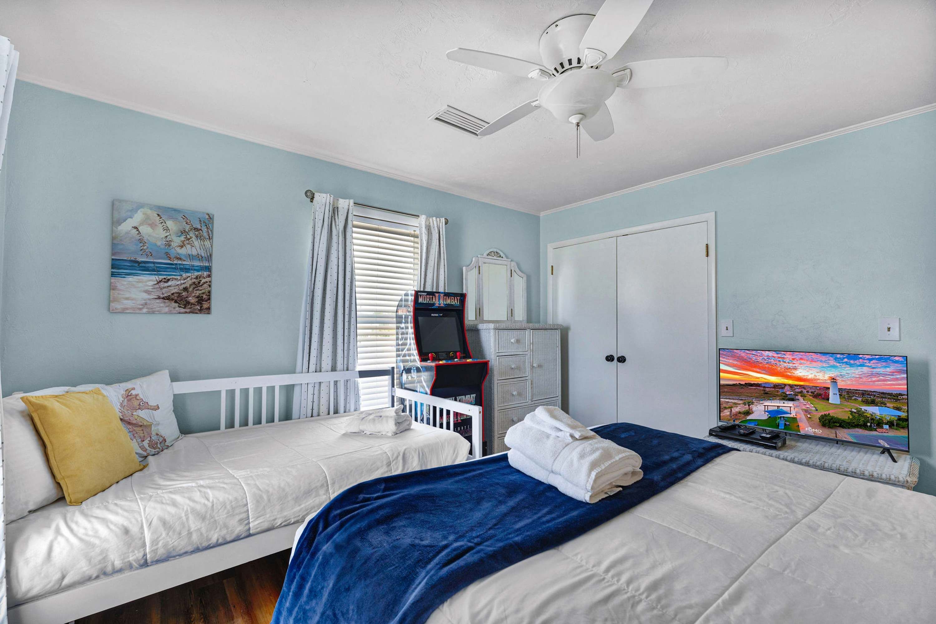Un dormitorio con dos camas, un ventilador de techo, un televisor, una cuna y un cuadro en la pared.