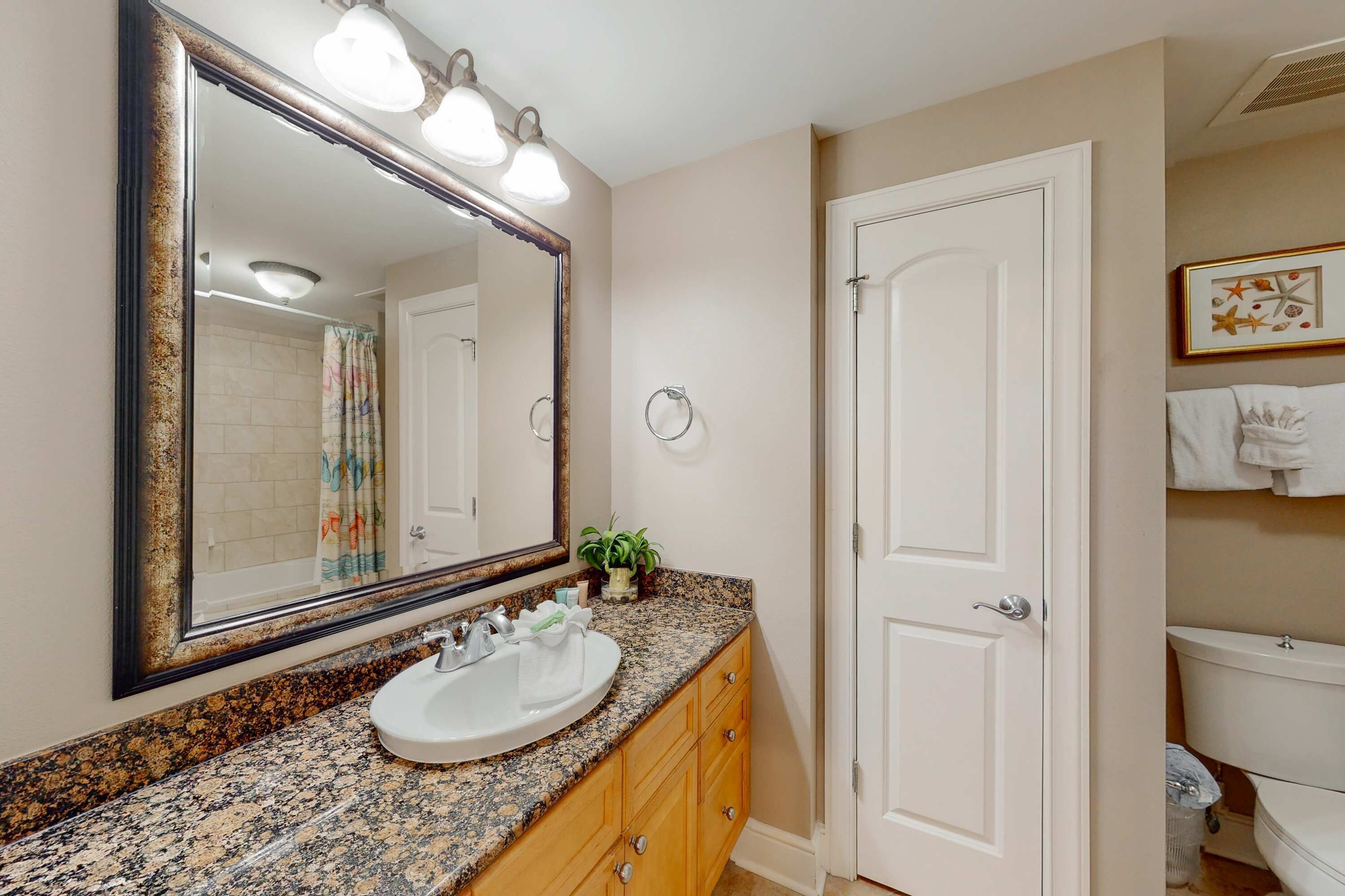 A bathroom with a granite countertop, a white sink, a large mirror with lights, wooden cabinets, and a white door.