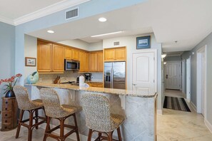 A kitchen with a central island, bar stools, wooden cabinets, stainless steel appliances, and a view into the adjacent dining area. - Beautiful coastal retreat with water view, beach access, & shared pool,hot tub (Fort Walton Beach)