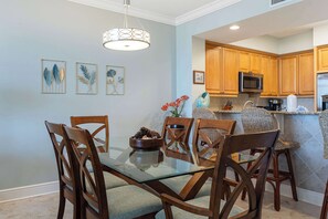 A dining area with a glass table and wooden chairs, a kitchen with wooden cabinets and stainless steel appliances, and wall art above the table. - Beautiful coastal retreat with water view, beach access, & shared pool,hot tub (Fort Walton Beach)