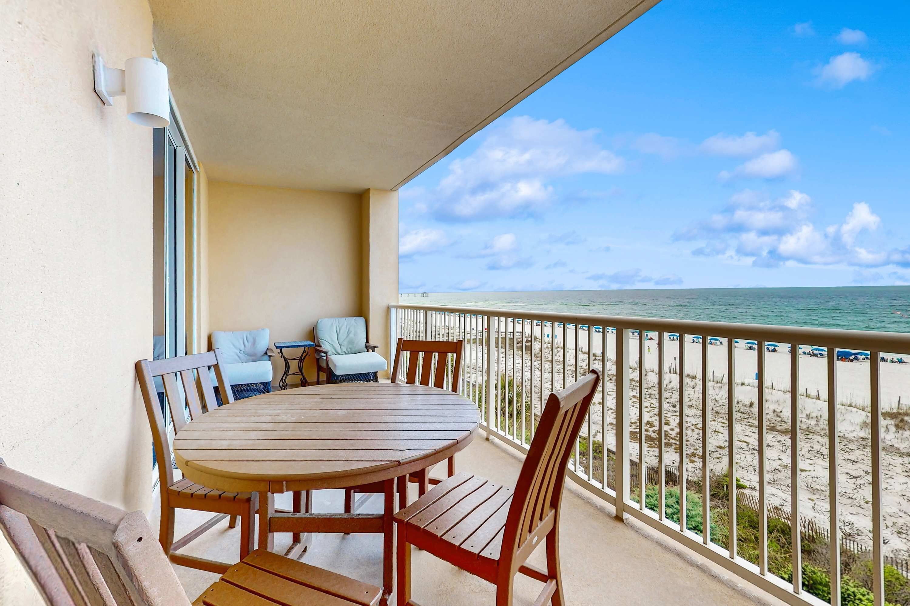 A balcony with a wooden table and chairs, overlooking a beach with a fence and a pier in the distance.