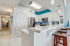 A modern kitchen with white cabinets, a stainless steel refrigerator, a microwave, and a large island with bar stools. - Gulf Front Condo, Beach Service Included, Walk To Dining & Entertainment (Fort Walton Beach)