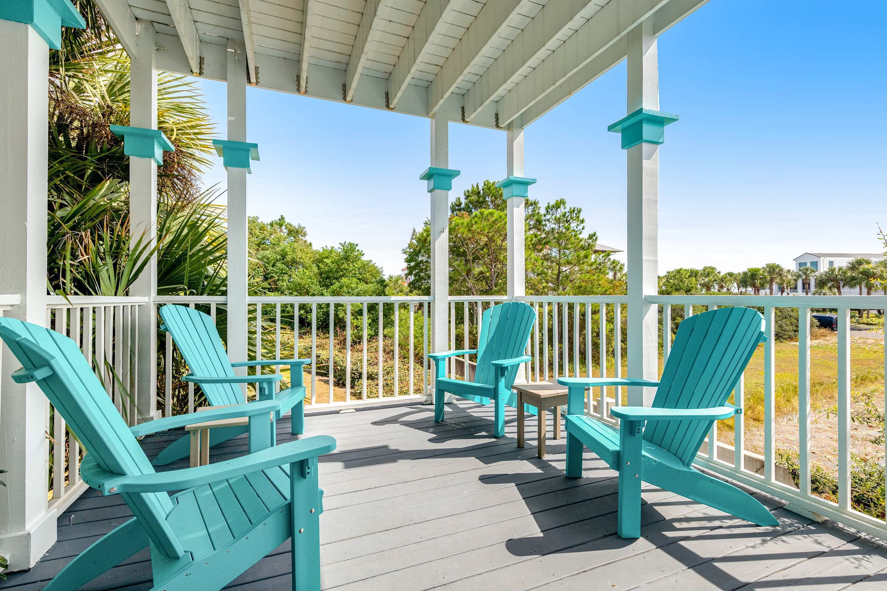 A covered porch with turquoise Adirondack chairs and a small table, overlooking a beach and greenery.