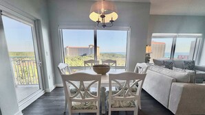 A spacious living room with a dining area, large windows with a view of a building and greenery, and a modern chandelier. - Gorgeous coastal condo with shared pool and private balcony (Miramar Beach)