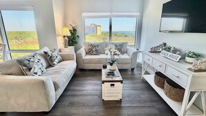 A modern living room with a beige sofa, a coffee table, and a TV on a white cabinet. - Gorgeous coastal condo with shared pool and private balcony (Miramar Beach)