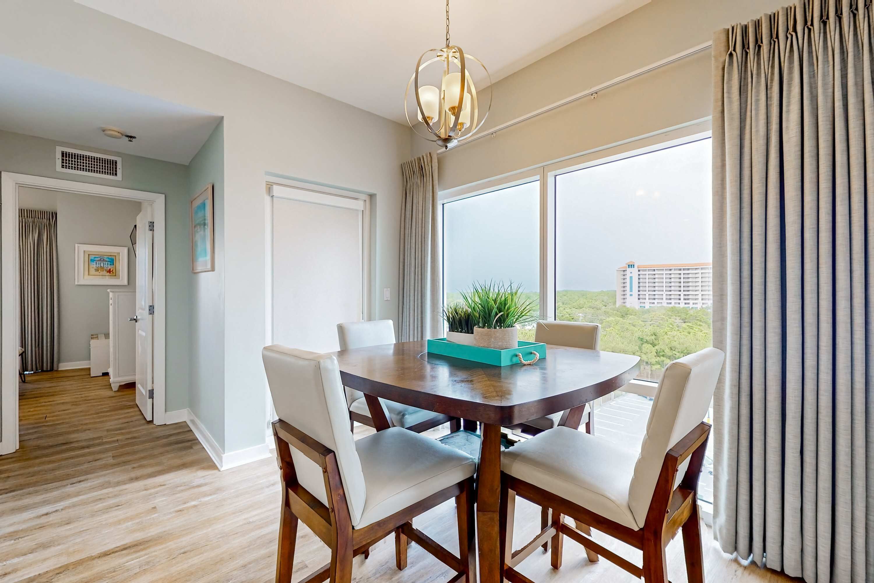 A dining area with a round table, four chairs, a chandelier, and a view of a building through large windows.