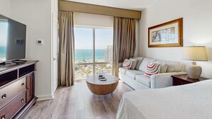 Habitació d'hotel amb finestra gran, televisió de pantalla plana, taula de centre rodona de fusta, sofà amb coixinets estampats i una làmpada.