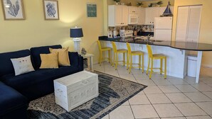 Una sala de estar con un sofá azul oscuro, un aparador blanco, un tapete estampado y una cocina con armarios blancos, una encimera negra y taburetes amarillos.