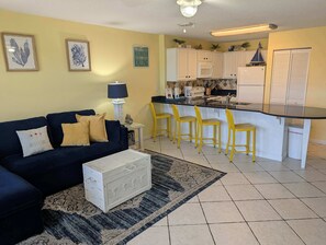 A living room with a dark blue sofa, a white chest of drawers, a patterned rug, and a kitchen with white cabinets, a black countertop, and yellow bar stools. - Beautiful seaside condo featuring a sparkling pool and direct beach access (Fort Walton Beach)