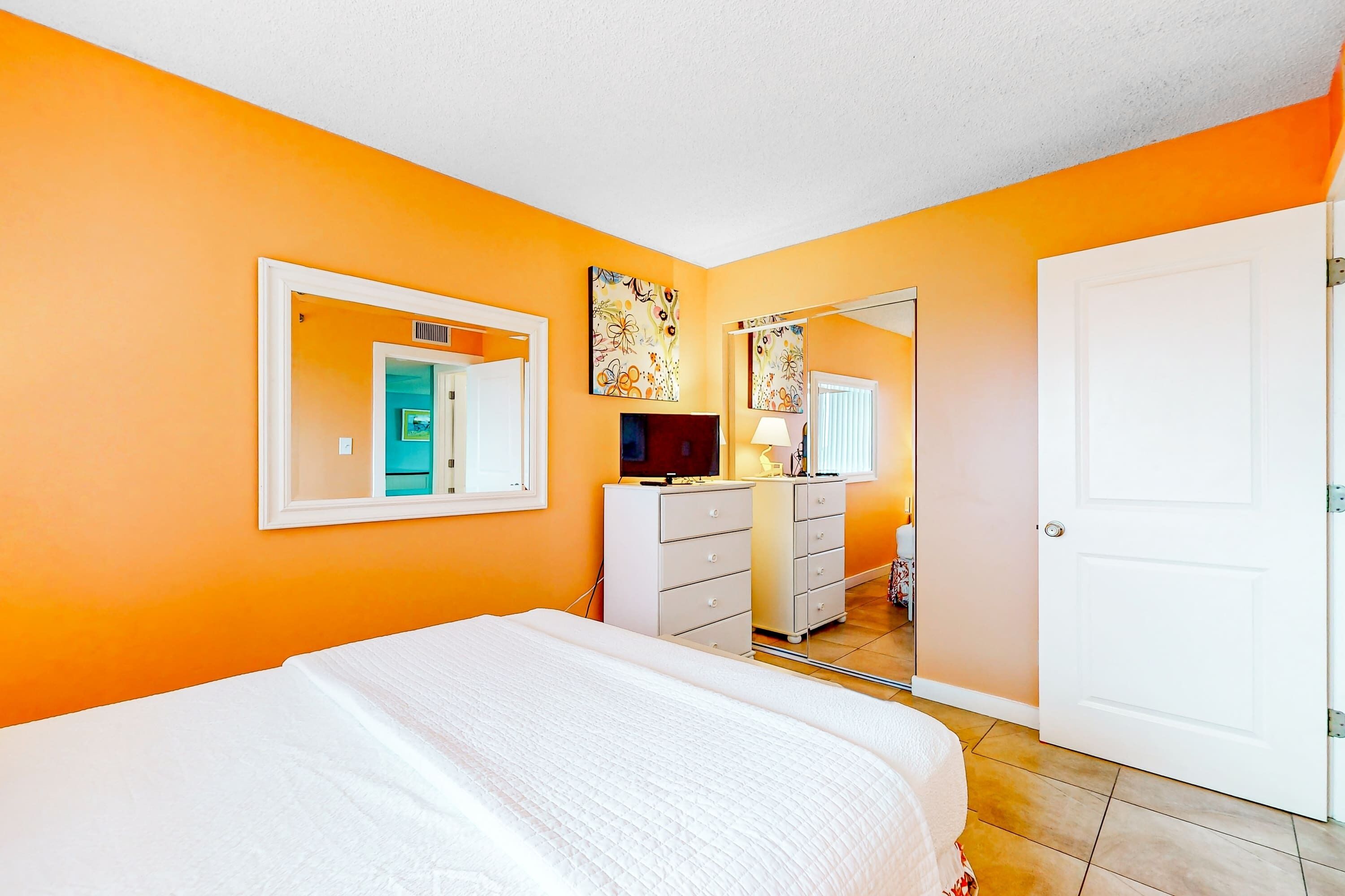 Une chambre avec des murs orange, un lit blanc, une commode, un miroir et une porte.