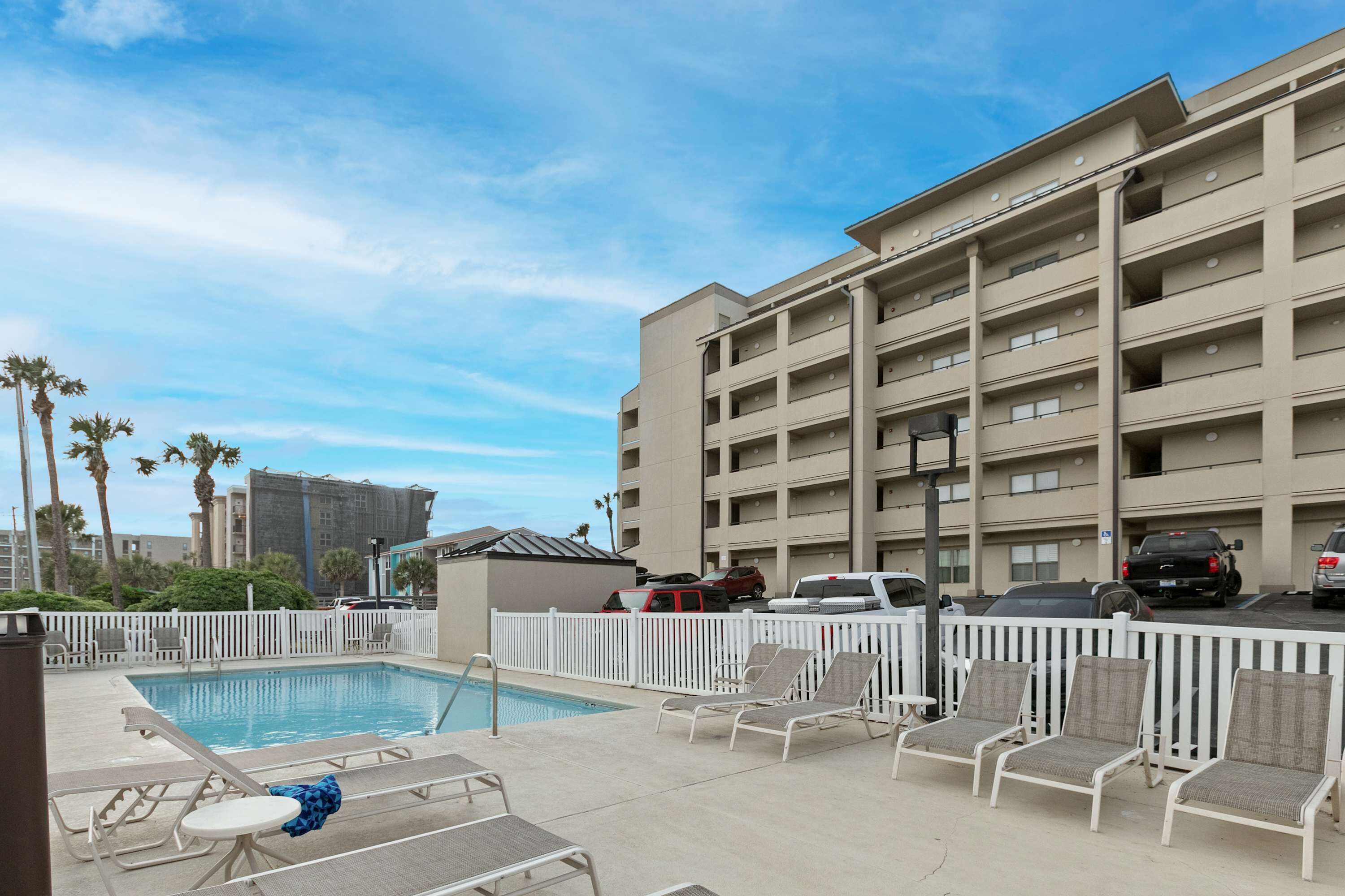 A hotel pool area with lounge chairs, a pool, and a parking lot with cars.