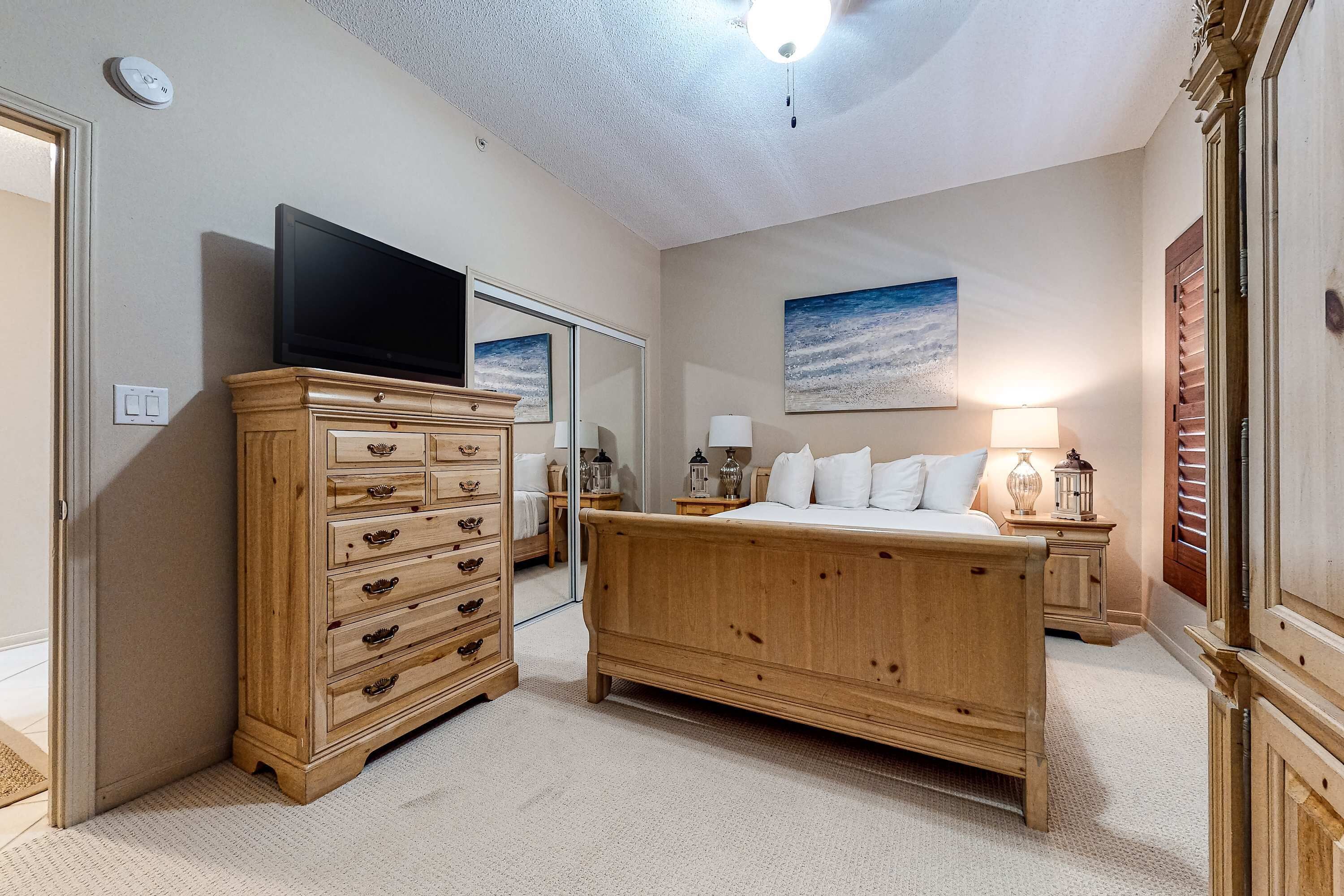 Ein Schlafzimmer mit einem hölzernen Schminktisch, einem Bett, einem Fernseher, einem Nachttisch und einem Gemälde an der Wand.
