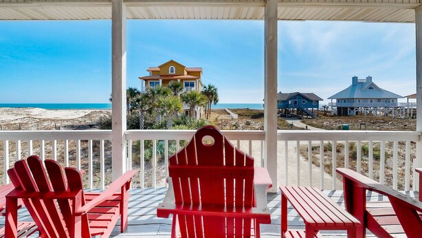 A covered porch with red Adirondack chairs and a table, overlooking a beach with houses and a clear sky. - Beachside home with private pool & amazing views (St. George Island)