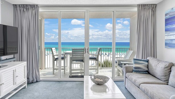 A modern hotel room with a balcony overlooking a beach, a flat-screen TV, a sofa, and a coffee table. - Seaside condo with wonderful beach views, balcony, grill, pool, hot tub, & gym (Fort Walton Beach)