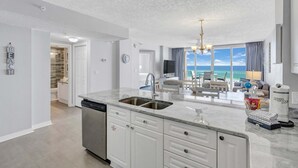 A modern kitchen with a large island, a dishwasher, and a sink. There is a dining area with a view of the ocean. - Seaside condo with wonderful beach views, balcony, grill, pool, hot tub, & gym (Fort Walton Beach)