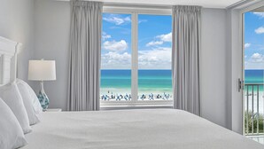 A hotel room with a large bed, a nightstand with a lamp, and a view of the beach through a large window. - Seaside condo with wonderful beach views, balcony, grill, pool, hot tub, & gym (Fort Walton Beach)