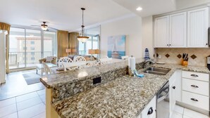 A modern kitchen with granite countertops, white cabinetry, and a dining area with white chairs and a table. - Floor Gulf Front Condo Fun Amenities, Nearby Shopping & Dining (Fort Walton Beach)
