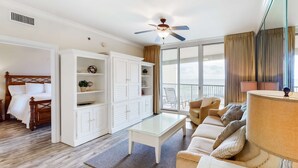 A modern living room with a glass coffee table, a sofa, a ceiling fan, and a large window with curtains. - Floor Gulf Front Condo Fun Amenities, Nearby Shopping & Dining (Fort Walton Beach)