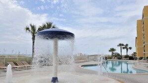 A pool area with a water feature in the center, surrounded by lounge chairs and palm trees. - Floor Gulf Front Condo Fun Amenities, Nearby Shopping & Dining (Fort Walton Beach)