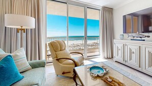 A spacious room with a large window offering a view of the beach, a comfortable sofa, a leather armchair, a television, and a cabinet. - Pretty Gulf Front Condo Fun Amenities, Nearby Shopping & Dining! (Fort Walton Beach)