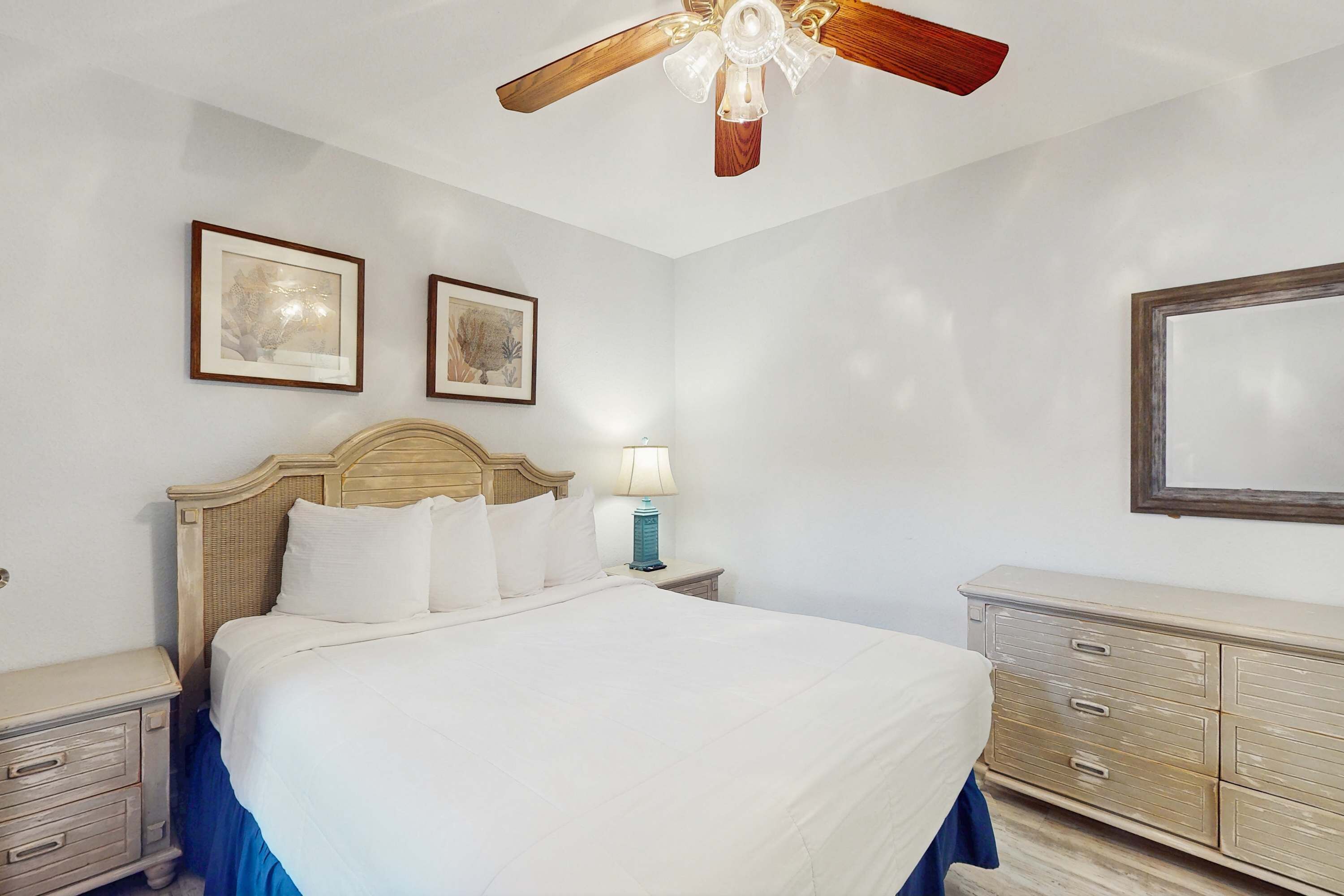 A hotel room with a large bed, a wooden headboard, a nightstand with a lamp, a dresser, and two framed pictures on the wall.