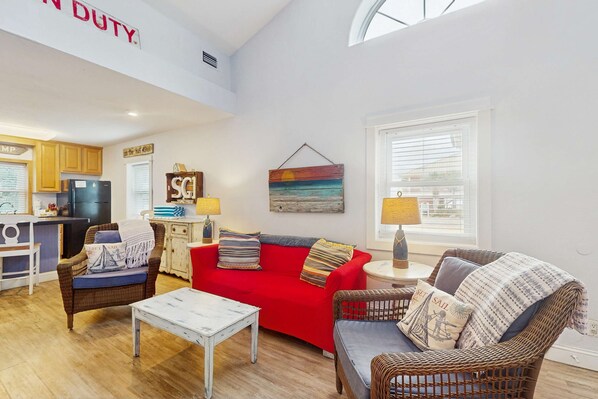 A spacious living area with a red sofa, wicker chairs, a small white table, and a kitchen in the background.