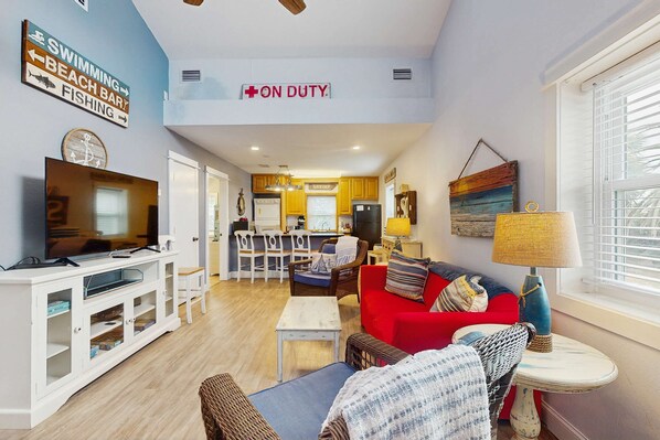 A living room with a TV, sofa, and dining area. There is a kitchen in the background with a sign that says "ON DUTY."