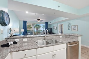 A modern kitchen with granite countertops, a stainless steel sink, and a dishwasher. There is a large window with a view of palm trees and a balcony. - Shores of Panama #216 1BR+Bunk, 2nd floor! (Panama City Beach)