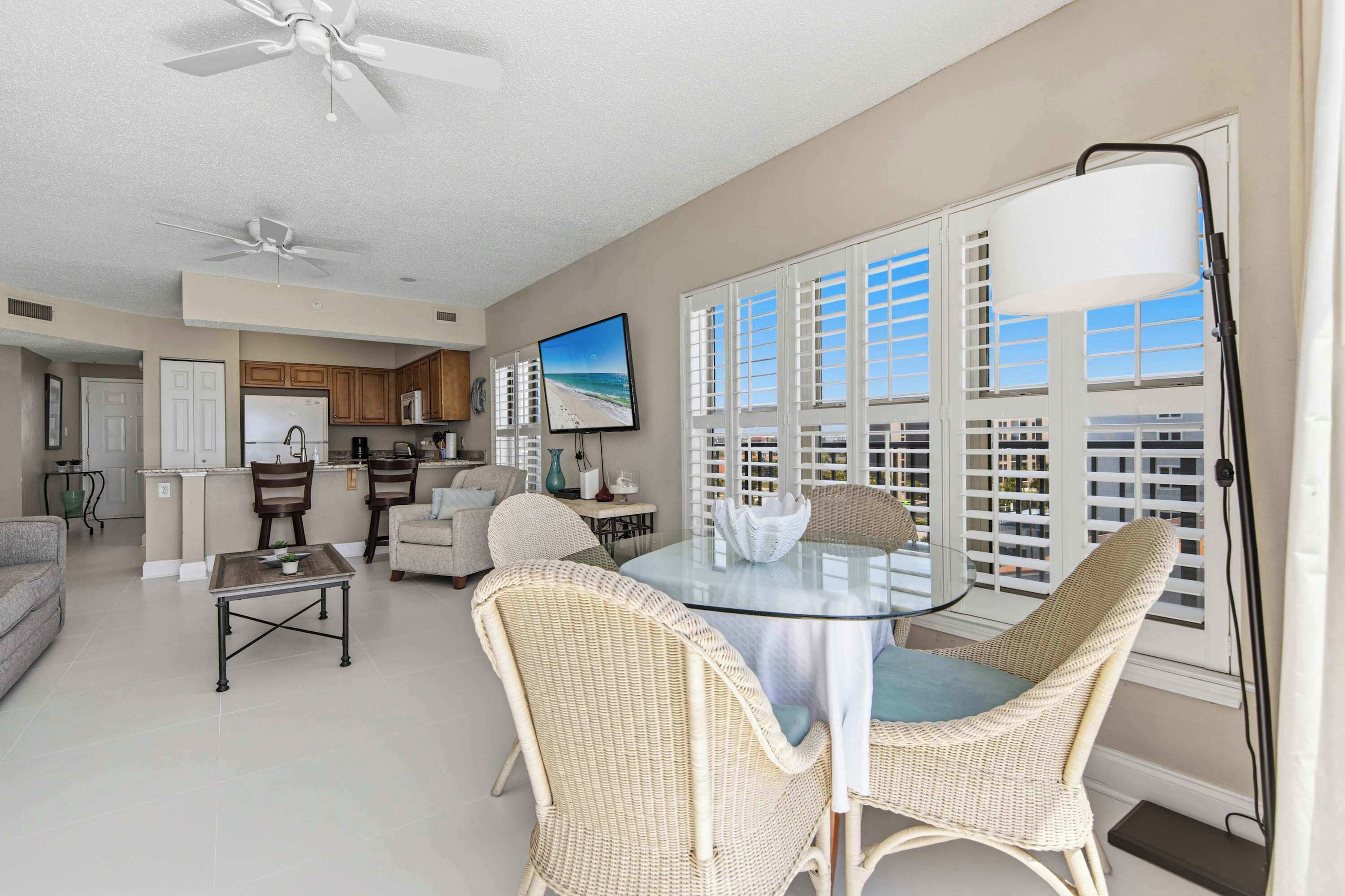 A spacious living area with a dining table, wicker chairs, a ceiling fan, a kitchen in the background, and a large window with shutters.
