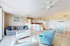 Ein großzügiger Wohnbereich mit Sofa, Sesseln, Couchtisch und Esstisch mit Stühlen. Es gibt eine Küchenzeile mit weißen Schränken und einem Kühlschrank. Ein Deckenventilator ist sichtbar, und ein Gemälde hängt an der Wand.