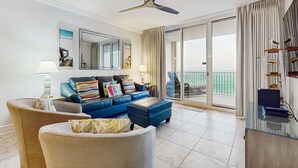 Ein geräumiges Wohnzimmer mit einem blauen Sofa, Korbstühlen, einem Glastisch und einem Deckenventilator.