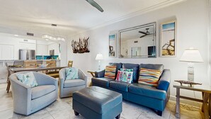 Ein modernes Wohnzimmer mit einer Couch, zwei Sesseln, einem Couchtisch, einem Essbereich mit Tisch und Stühlen, einer Küche mit Geräten und einem Deckenventilator.