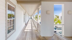 Un corredor cubierto con paredes blancas y piso de madera, que conduce a un balcón con vistas a coches aparcados y palmeras.